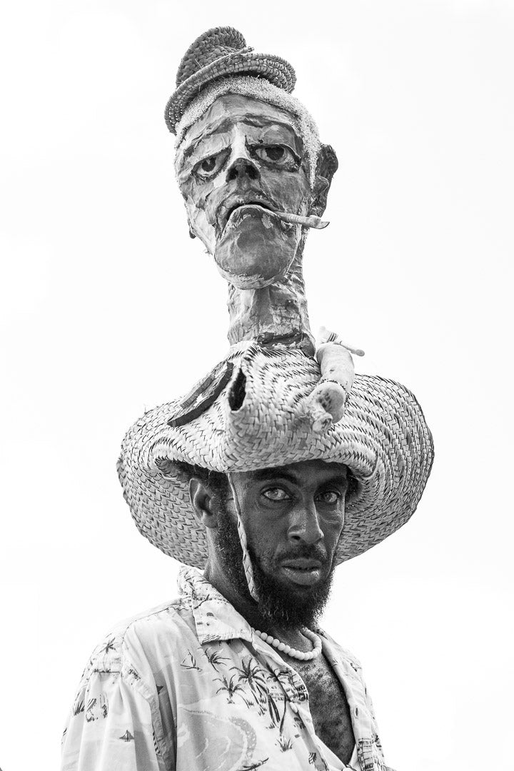 Person wearing a large decorative hat with a mask-like face on top against a white background